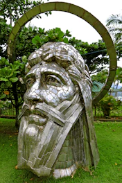 A large metallic sculpture of a face surrounded by lush greenery, featuring detailed facial features and a halo above the head. The surrounding garden includes various trees and shrubs, creating a harmonious natural setting.