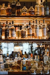 Multiple bottles of whiskey and spirits are displayed on a wooden shelf with warm lighting that highlights the labels. The scene appears to be in a bar setting with a luxurious and sophisticated atmosphere. The background shows a blurred view of people seated, adding to the ambience.