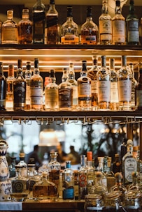 Multiple bottles of whiskey and spirits are displayed on a wooden shelf with warm lighting that highlights the labels. The scene appears to be in a bar setting with a luxurious and sophisticated atmosphere. The background shows a blurred view of people seated, adding to the ambience.