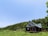 A small wooden cabin is nestled in a lush green field surrounded by dense forest. The sky is clear and blue, casting a serene and tranquil atmosphere. The cabin has a slanted roof, large windows, and a porch, blending harmoniously with the natural surroundings.