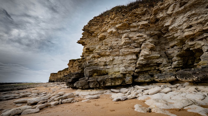 A rugged cliff face made of layered rocks stands prominently against a cloudy sky. The foreground features a sandy beach scattered with large, smooth stones. The texture of the rock layers highlights the geological formation, while the overcast sky adds a dramatic backdrop.