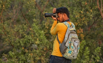 A person is standing outdoors in a lush, green setting, focusing intently while looking through a camera. They are wearing a yellow shirt and a colorful backpack, blending in with the natural environment around them.