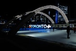 An urban scene at night featuring an illuminated sign spelling 'Toronto' with decorative arches and stars overhead. A skating rink occupies the foreground, with figures skating against the backdrop of city buildings and lights.