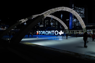 An urban scene at night featuring an illuminated sign spelling 'Toronto' with decorative arches and stars overhead. A skating rink occupies the foreground, with figures skating against the backdrop of city buildings and lights.