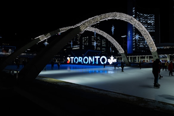 An urban scene at night featuring an illuminated sign spelling 'Toronto' with decorative arches and stars overhead. A skating rink occupies the foreground, with figures skating against the backdrop of city buildings and lights.