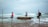 Three people are fishing while seated on stilt poles in shallow ocean water. The ocean waves crash gently around them, and they each hold a fishing rod. The sky above is overcast, creating a tranquil and serene setting.