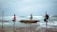 Three people are fishing while seated on stilt poles in shallow ocean water. The ocean waves crash gently around them, and they each hold a fishing rod. The sky above is overcast, creating a tranquil and serene setting.