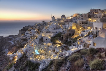 A picturesque coastal village with whitewashed buildings, domed roofs, and narrow winding streets, illuminated by warm lights at dusk. The sea extends into the horizon, and a windmill is visible amidst the architecture perched on a hillside.