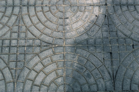 Grey paving stones arranged in a circular pattern with intersecting lines creating geometric shapes. The surface has a slightly weathered appearance, with light and shadow variations on the textured surface.