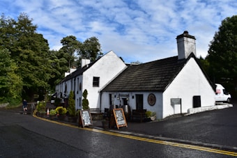 A rustic white building with a steep dark roof, likely a pub or inn, is set against a backdrop of lush green trees. The facade includes several small windows and a chimney. Wooden barrels and chalkboard signs advertising food and live music are placed by the entrance. A paved road with yellow lines runs alongside, leading to a parked white van by the building. Puffy clouds fill the bright blue sky.