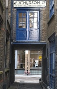 A narrow alleyway features an old brick building with a sign for 'W. A. Hudson Ltd Furnishing Brassfounders'. A blue door and window above an archway contrasts with the surrounding brickwork. Through the passage, a pink bicycle rests against a modern building with large glass windows.