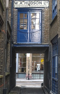 A narrow alleyway features an old brick building with a sign for 'W. A. Hudson Ltd Furnishing Brassfounders'. A blue door and window above an archway contrasts with the surrounding brickwork. Through the passage, a pink bicycle rests against a modern building with large glass windows.
