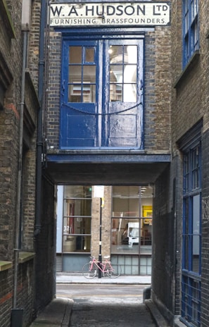 A narrow alleyway features an old brick building with a sign for 'W. A. Hudson Ltd Furnishing Brassfounders'. A blue door and window above an archway contrasts with the surrounding brickwork. Through the passage, a pink bicycle rests against a modern building with large glass windows.