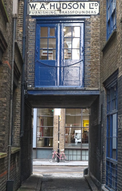 A narrow alleyway features an old brick building with a sign for 'W. A. Hudson Ltd Furnishing Brassfounders'. A blue door and window above an archway contrasts with the surrounding brickwork. Through the passage, a pink bicycle rests against a modern building with large glass windows.