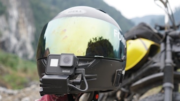 A motorcycle helmet with a reflective visor mounted with a camera is prominently displayed. The helmet is positioned next to a yellow motorcycle in a scenic, mountainous landscape.