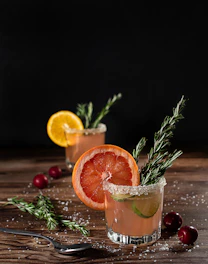 A vibrant cocktail glass garnished with fresh herbs and citrus on a wooden table with a Jura landscape background.