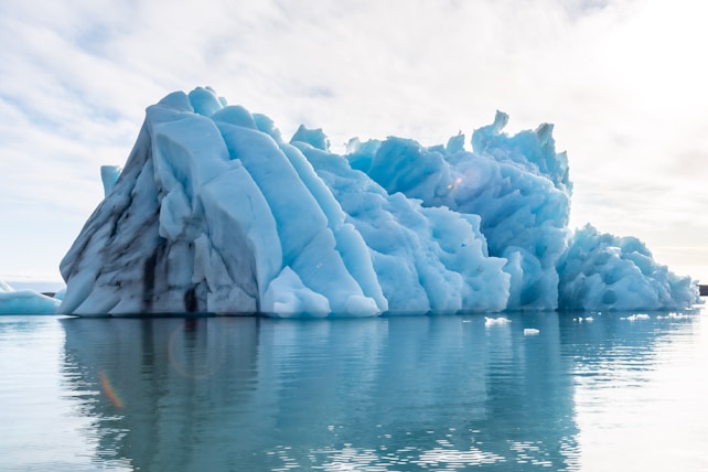 A massive iceberg with jagged, towering formations floats serenely on calm waters. The iceberg's surface is a striking blend of brilliant blue and white ice, with dark streaks and intricate textures visible. The sky is overcast with a soft, diffused light that casts reflections on the water, enhancing the serene and majestic atmosphere.