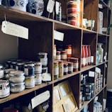 A wooden shelf filled with various jars, bottles, and packages of food products, including truffles, sauces, pasta, and preserved goods. Each shelf holds differently sized containers with labels and tags, creating a rustic and artisanal ambiance.