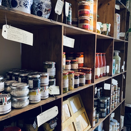 A wooden shelf filled with various jars, bottles, and packages of food products, including truffles, sauces, pasta, and preserved goods. Each shelf holds differently sized containers with labels and tags, creating a rustic and artisanal ambiance.