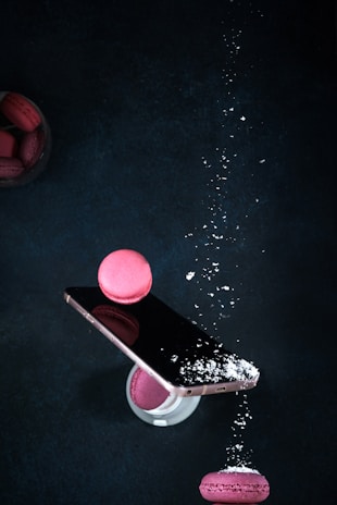 A smartphone balanced at an angle, topped with a pink macaron, and sprinkled with powdered sugar. Additional macarons are seen around the phone, with one on a glass and another beside the phone, against a dark background.