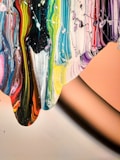 Vibrant swirls of paint cascade across the canvas, featuring a spectrum of colors including blues, greens, yellows, reds, and pinks. The paint appears thick and textured, partially blending yet maintaining distinct lines of color. A smooth, peach-toned background contrasts with the vivid colors.
