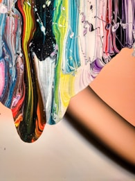 Vibrant swirls of paint cascade across the canvas, featuring a spectrum of colors including blues, greens, yellows, reds, and pinks. The paint appears thick and textured, partially blending yet maintaining distinct lines of color. A smooth, peach-toned background contrasts with the vivid colors.