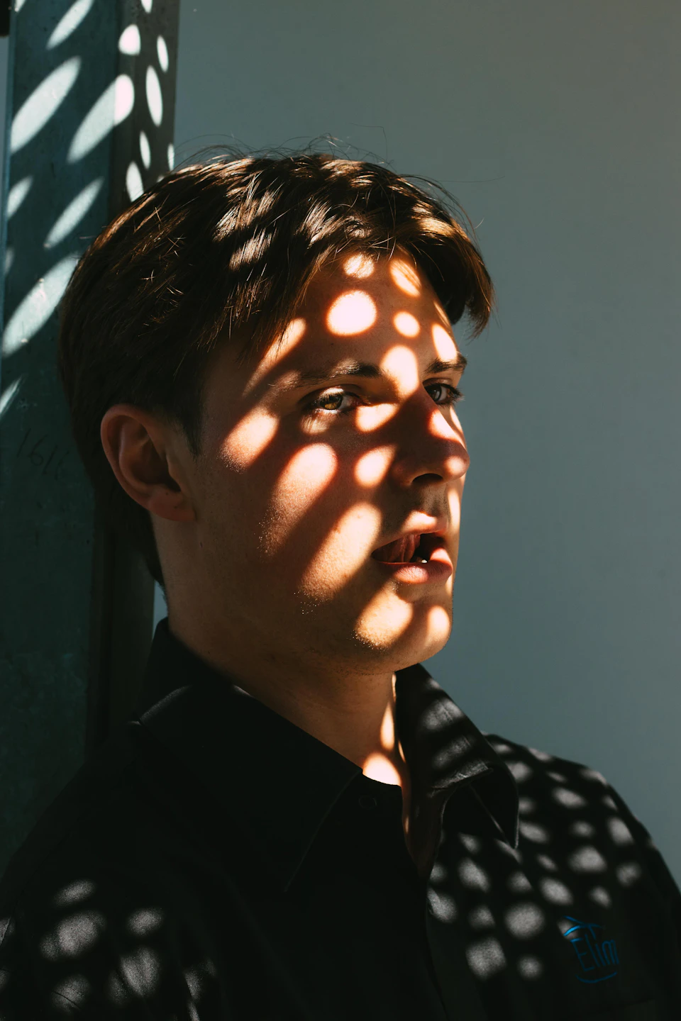 Young man with short brown hair stands against white wall, illuminated by sunlight streaming through