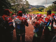 A group of people wearing colorful, matching outfits gather outdoors, appearing to be a part of a parade or festival. They are playing musical instruments, with some holding drums and other percussion items. The scene is lively and surrounded by trees with a large rock formation visible in the background.