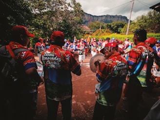 A group of people wearing colorful, matching outfits gather outdoors, appearing to be a part of a parade or festival. They are playing musical instruments, with some holding drums and other percussion items. The scene is lively and surrounded by trees with a large rock formation visible in the background.