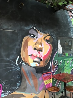 A vibrant mural on a dark wall depicts a stylized portrait of a person with an afro hairstyle. The face is painted with a mix of warm and cool colors, including shades of brown, pink, and orange. Piercing eyes and full lips are prominent features. Below the mural, two wooden tables with pink and yellow chairs add a contrasting touch.