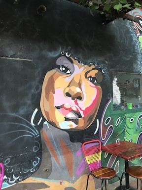A vibrant mural on a dark wall depicts a stylized portrait of a person with an afro hairstyle. The face is painted with a mix of warm and cool colors, including shades of brown, pink, and orange. Piercing eyes and full lips are prominent features. Below the mural, two wooden tables with pink and yellow chairs add a contrasting touch.