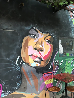 A vibrant mural on a dark wall depicts a stylized portrait of a person with an afro hairstyle. The face is painted with a mix of warm and cool colors, including shades of brown, pink, and orange. Piercing eyes and full lips are prominent features. Below the mural, two wooden tables with pink and yellow chairs add a contrasting touch.