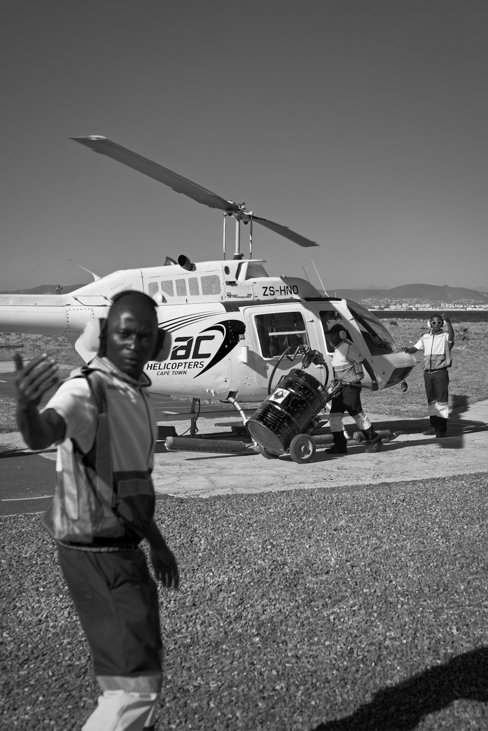 Foto en escala de grises de un hombre con camisa y pantalones grises de pie cerca de un helicóptero blanco y rojo