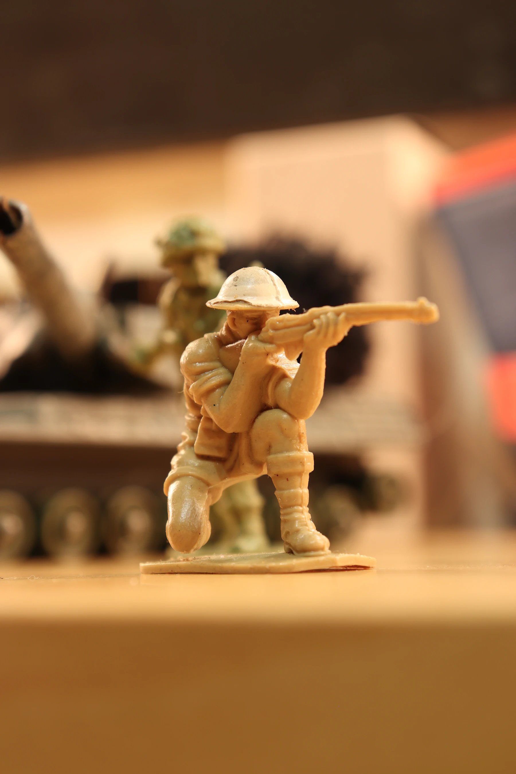 An unpainted tan plastic soldier kneeling and aiming a rifle, with a model tank blurred behind
