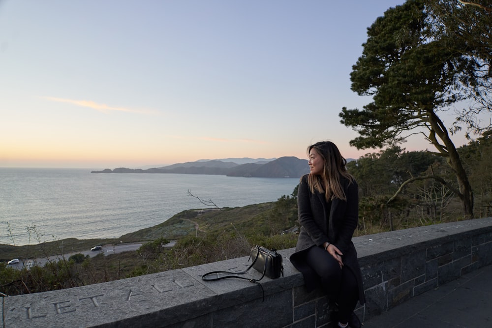 woman in black jacket sitting on gray concrete bench near body of water during daytime