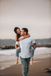 man in white crew neck t-shirt kissing woman in blue denim jeans on beach during