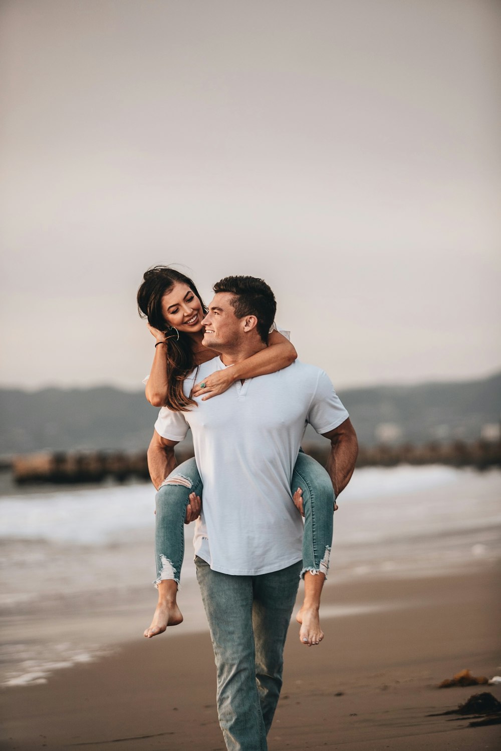 man in white crew neck t-shirt kissing woman in blue denim jeans on beach during