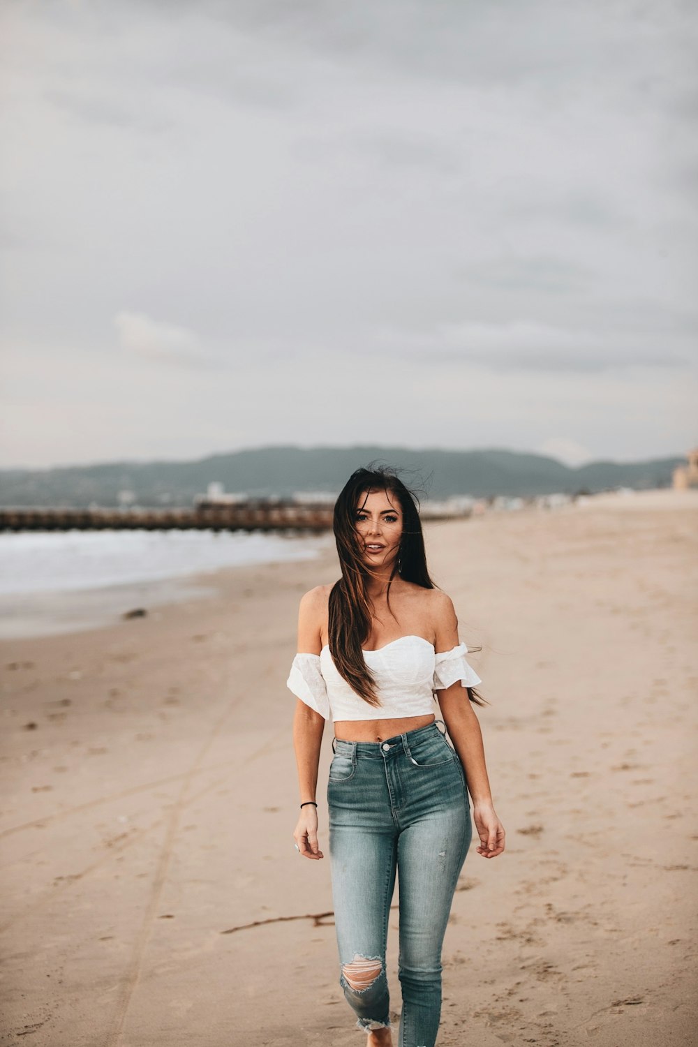 woman in white off shoulder shirt and blue denim jeans standing on beach shore during daytime