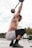 A man is engaged in an overhead squat exercise with a barbell. His body is adorned with multiple tattoos, and he is wearing colorful shorts and black shoes. He is in an outdoor gym setting with gym equipment visible in the background.
