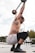 A man is engaged in an overhead squat exercise with a barbell. His body is adorned with multiple tattoos, and he is wearing colorful shorts and black shoes. He is in an outdoor gym setting with gym equipment visible in the background.