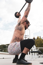 A man is engaged in an overhead squat exercise with a barbell. His body is adorned with multiple tattoos, and he is wearing colorful shorts and black shoes. He is in an outdoor gym setting with gym equipment visible in the background.