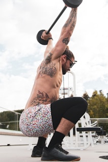 A man is engaged in an overhead squat exercise with a barbell. His body is adorned with multiple tattoos, and he is wearing colorful shorts and black shoes. He is in an outdoor gym setting with gym equipment visible in the background.