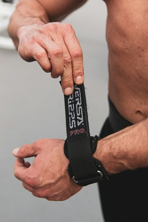 A muscular person adjusting a black wrist strap with the text 'VERSA GRIP PRO' on it. The focus is on the hands and forearms, showcasing the details of the strap. The background is neutral, emphasizing the subject.