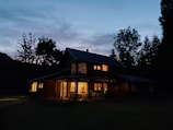 Evening shot of a rustic yet modern country house illuminated warmly against a twilight backdrop.