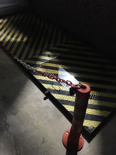 A dimly lit area features a traffic cone with white reflective stripes, connected by a red and white chain to another point. The ground has a textured surface with yellow and black chevron stripes, possibly indicating a caution or restricted zone.