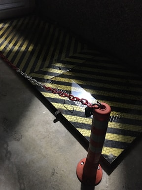 A dimly lit area features a traffic cone with white reflective stripes, connected by a red and white chain to another point. The ground has a textured surface with yellow and black chevron stripes, possibly indicating a caution or restricted zone.