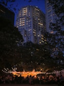 An evening scene features a brightly lit outdoor gathering beneath trees adorned with string lights. In the background, a tall building with numerous illuminated windows towers under a deep blue sky. People are seated and mingling under a tent, contributing to a lively yet relaxed atmosphere.