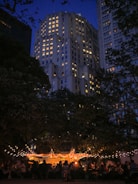 An evening scene features a brightly lit outdoor gathering beneath trees adorned with string lights. In the background, a tall building with numerous illuminated windows towers under a deep blue sky. People are seated and mingling under a tent, contributing to a lively yet relaxed atmosphere.