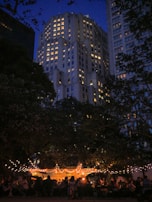 An evening scene features a brightly lit outdoor gathering beneath trees adorned with string lights. In the background, a tall building with numerous illuminated windows towers under a deep blue sky. People are seated and mingling under a tent, contributing to a lively yet relaxed atmosphere.