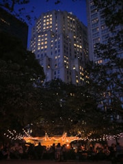 An evening scene features a brightly lit outdoor gathering beneath trees adorned with string lights. In the background, a tall building with numerous illuminated windows towers under a deep blue sky. People are seated and mingling under a tent, contributing to a lively yet relaxed atmosphere.